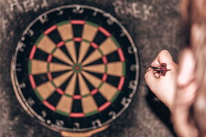 Im Vordergrund hält eine Person einen Darts-Pfeil in Wurfhaltung, im Hintergrund ist eine Dartscheibe an der Wand zu sehen.