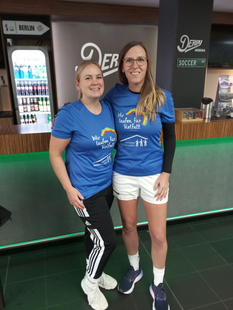 Die Geschäftsleitung, Ann Jerusalem, und die Gruppenleitung der Fussball-Gruppe, Caro Hoven, stehen Arm in Arm am Spielfeldrand. Beide tragen das blaue T-Shirt des Aachener Betreuungsbüros mit einem Regenbogen und der Aufschrift "Wir laufen für Vielfalt".