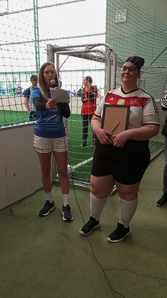 Die Geschäftsleitung, Ann Jerusalem, steht mit Mikrofon und Zetteln in der Hand neben dem Spielfeld. Eine Leistungsnehmende steht neben ihr, in Fussballkleidung, mit einem Bilderrahmen in der Hand.