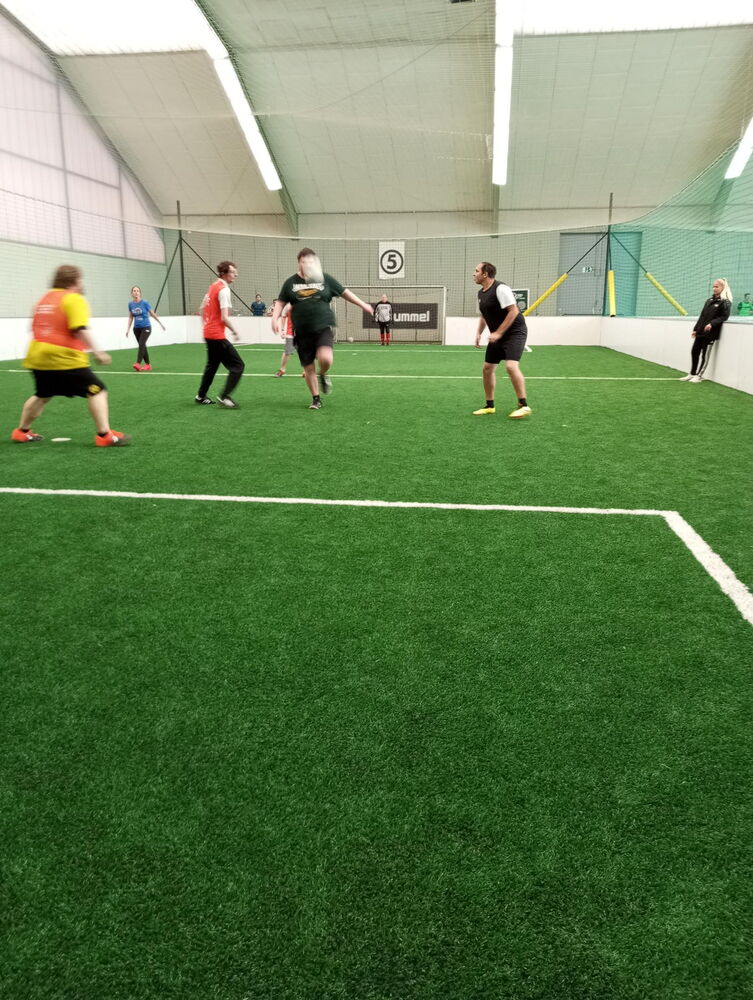 Vom Tor aus fotografiert sind die Spielenden zu sehen, der Ball befindet sich in der Luft. Der Boden ist saftig grüner Kunstrasen, das Spielfeld ist mit einem Netz umrundet.