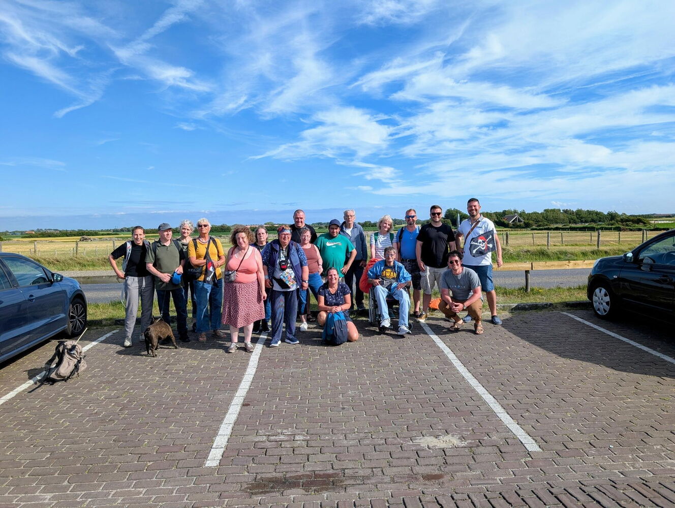 Insgesamt 13 Menschen, einer im Rollstuhl, die freudestrahlend in die Kamera blicken. Im Hintergrund ist blauer Himmel und gelb-grün blühende Landschaft.