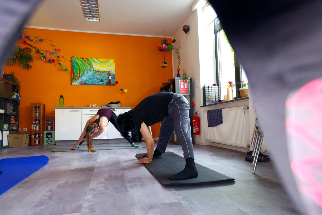 In einem Raum mit orangener Wand und buntem Bild sind drei Gymnastikmatten und zwei Personen zu sehen. Sie stehen mit gespreizten Beinen, die Hände auf dem Boden und dehnen sich zur rechten Seite.