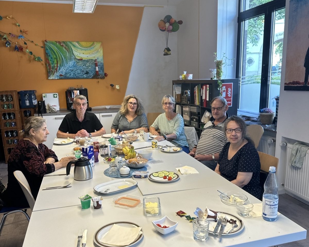 Gemeinsames Frühstück in einem Gemeinschaftsraum: Mehrere erwachsene Personen sitzen an einem großen Tisch mit Speisen und Getränken.