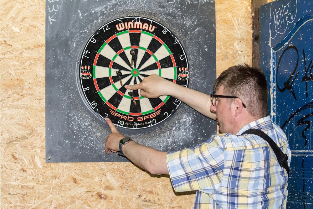 Beim Dartspiel zeigt eine Person auf die Trefferpositionen der Darts-Pfeile in der Dartscheibe.