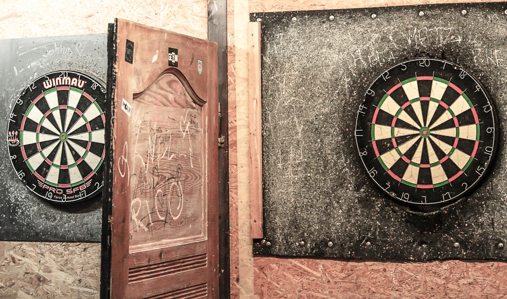 An einer Wand sind zwei Dartscheiben befestigt. Zwischen den Dartscheiben befindet sich eine Holztür mit Gebrauchsspuren als Abtrennung der Spielflächen.